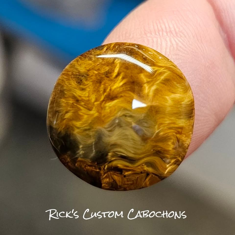 A pietersite cabochon held between two fingers, gold and amber fibre